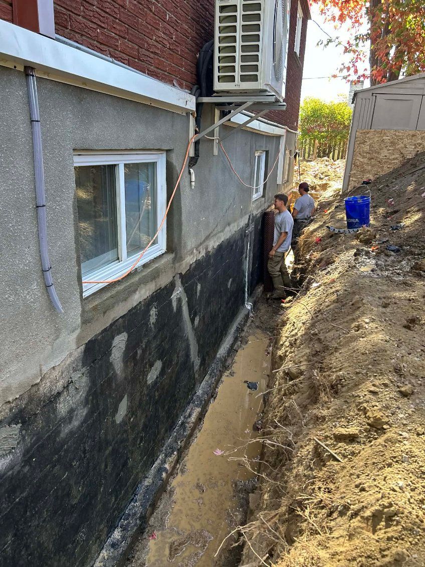 Des ouvriers effectuent des travaux d'excavation à proximité d'un bâtiment doté d'une membrane d'étanchéité noire nouvellement appliquée ; journée ensoleillée.