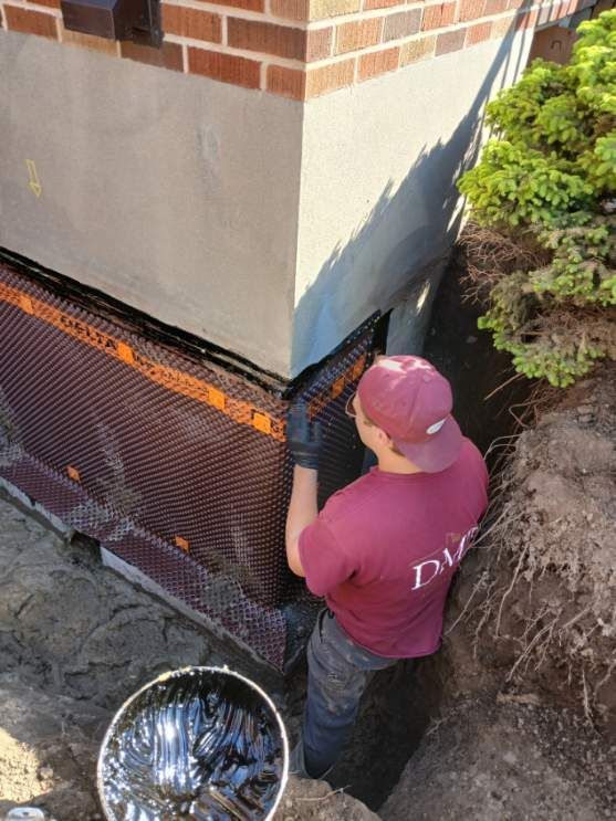 Un homme en chemise rouge installe une membrane de drainage de fondation à l'angle d'un bâtiment. On aperçoit de la terre, des briques et un seau.