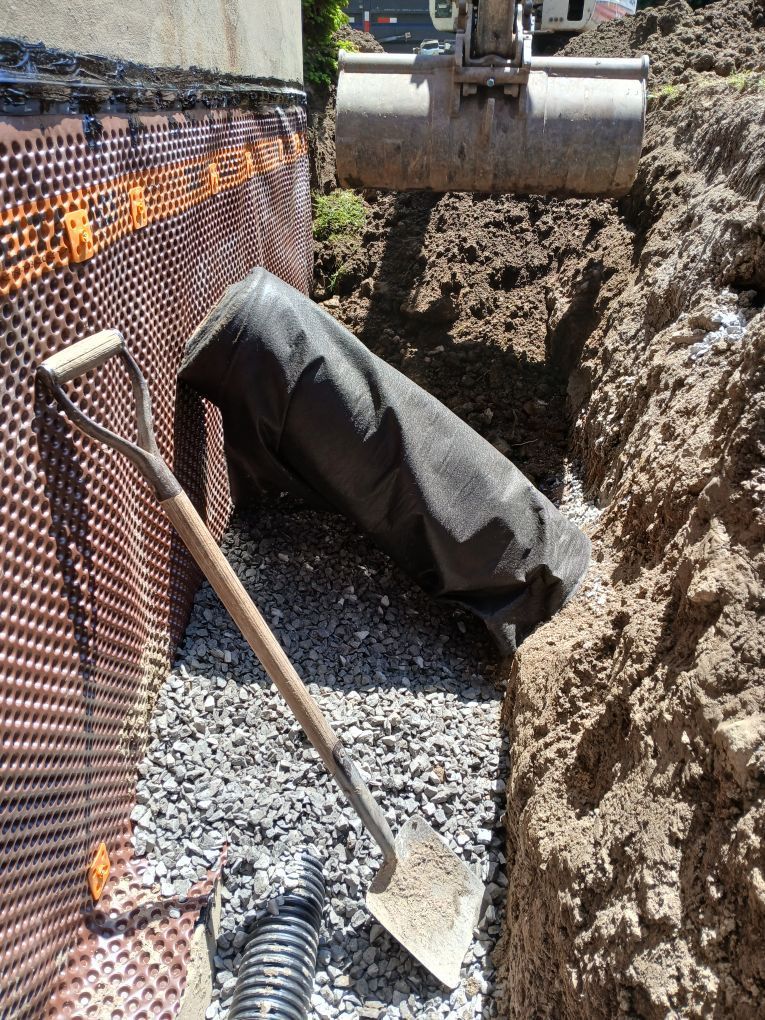 Membrane de drainage à picots noirs et orange installée dans un angle de fondation, entourée de terre et de briques.