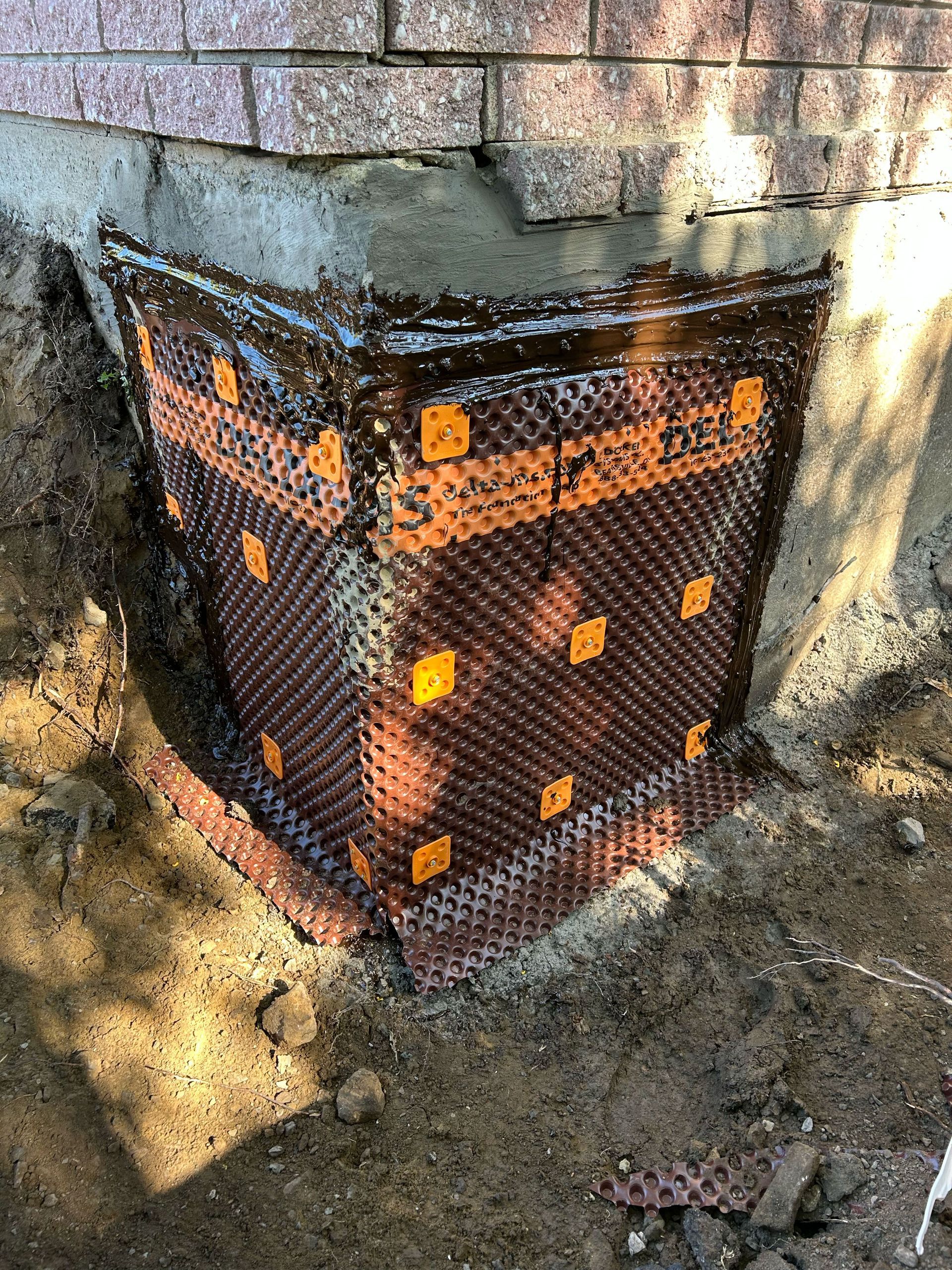 Membrane de drainage brune à picots enroulée autour d'un angle de fondation en briques, installée dans une tranchée.