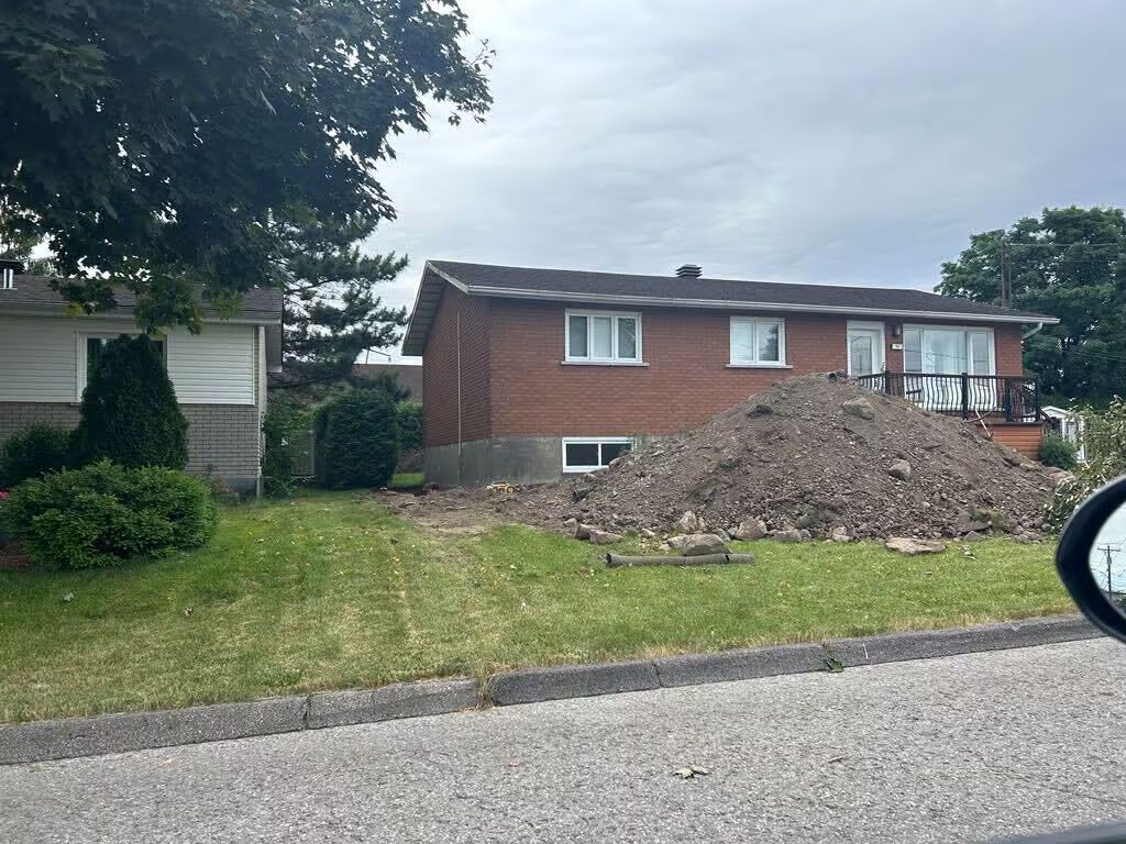 Une maison en briques de deux étages avec un grand tas de terre dans la cour avant. Ciel couvert.