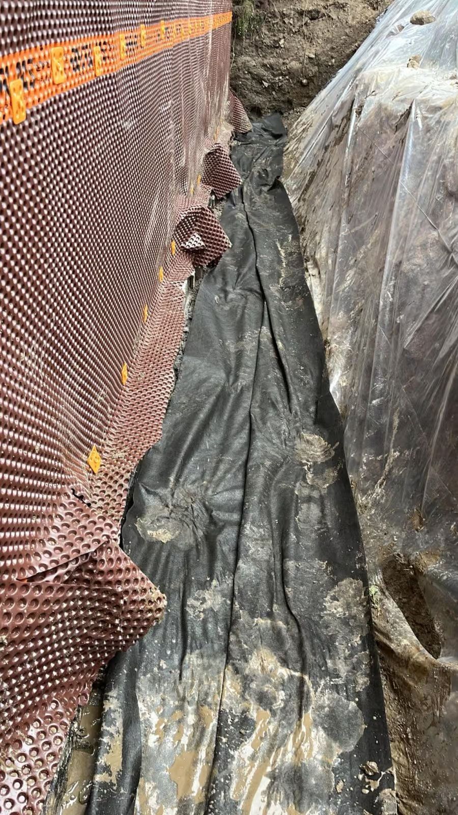 Mur de fondation brun avec tapis de drainage, tissu noir et terre dans une tranchée étroite.