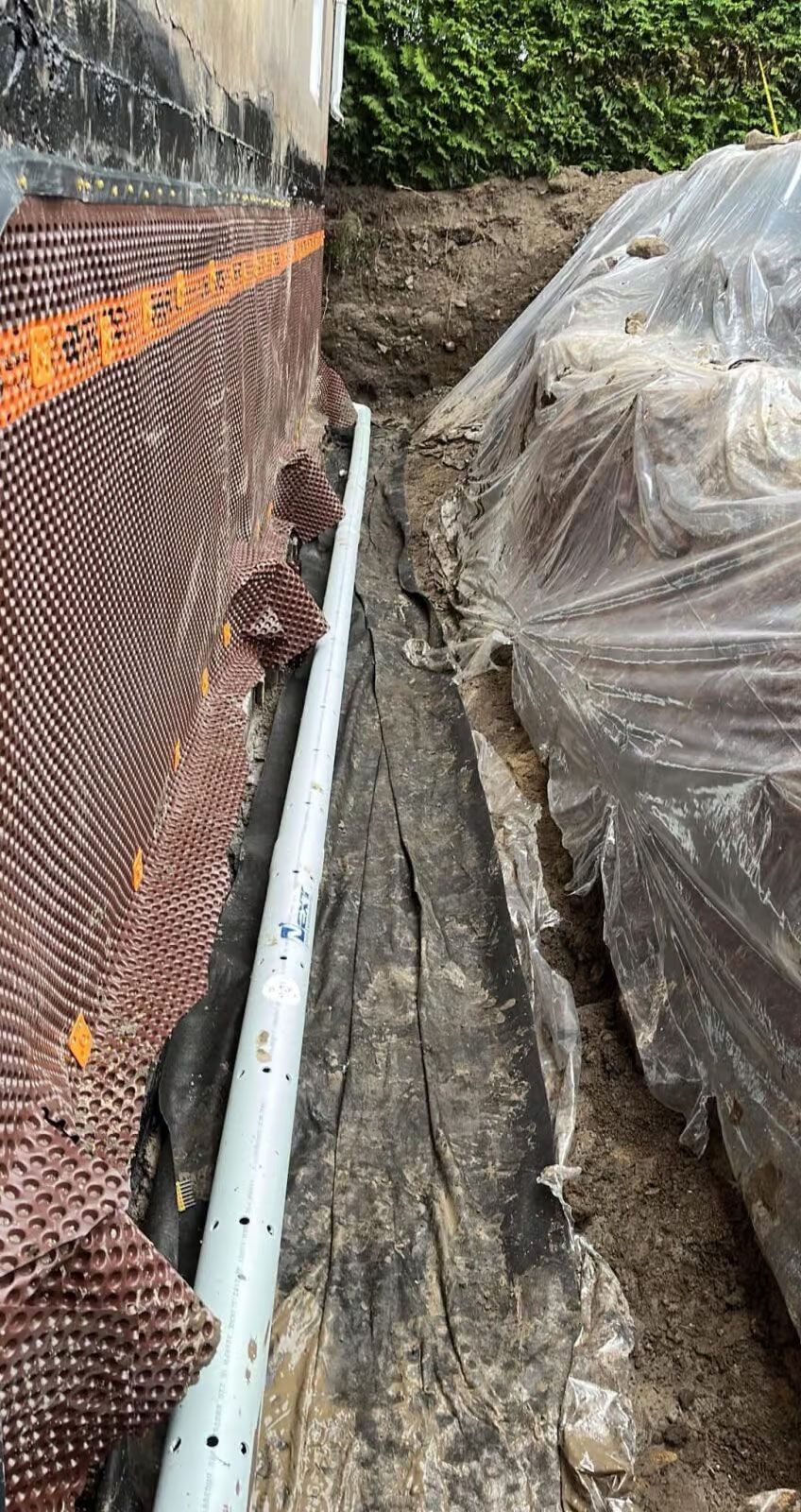 Un tuyau de drainage blanc perforé, installé à côté d'un mur de fondation recouvert d'une membrane de protection et d'un mur en pierre.