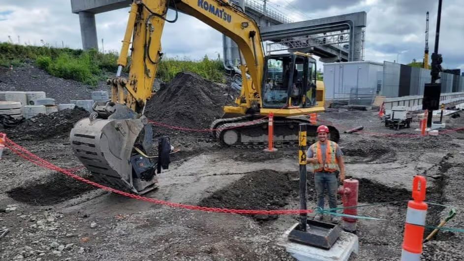 Chantier : Pelle mécanique et ouvrier inspectant le terrain, ruban de sécurité rouge, ciel couvert.