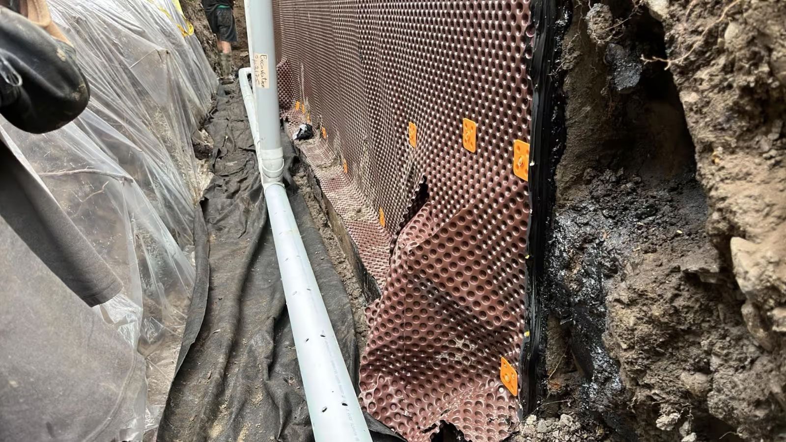 Installation d'un système de drainage dans une tranchée ; membrane brune à picots fixée au mur, tuyau blanc perforé.