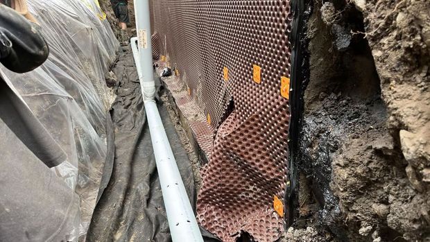 Installation d'un système de drainage dans une tranchée ; membrane brune à picots fixée au mur, tuyau blanc perforé.