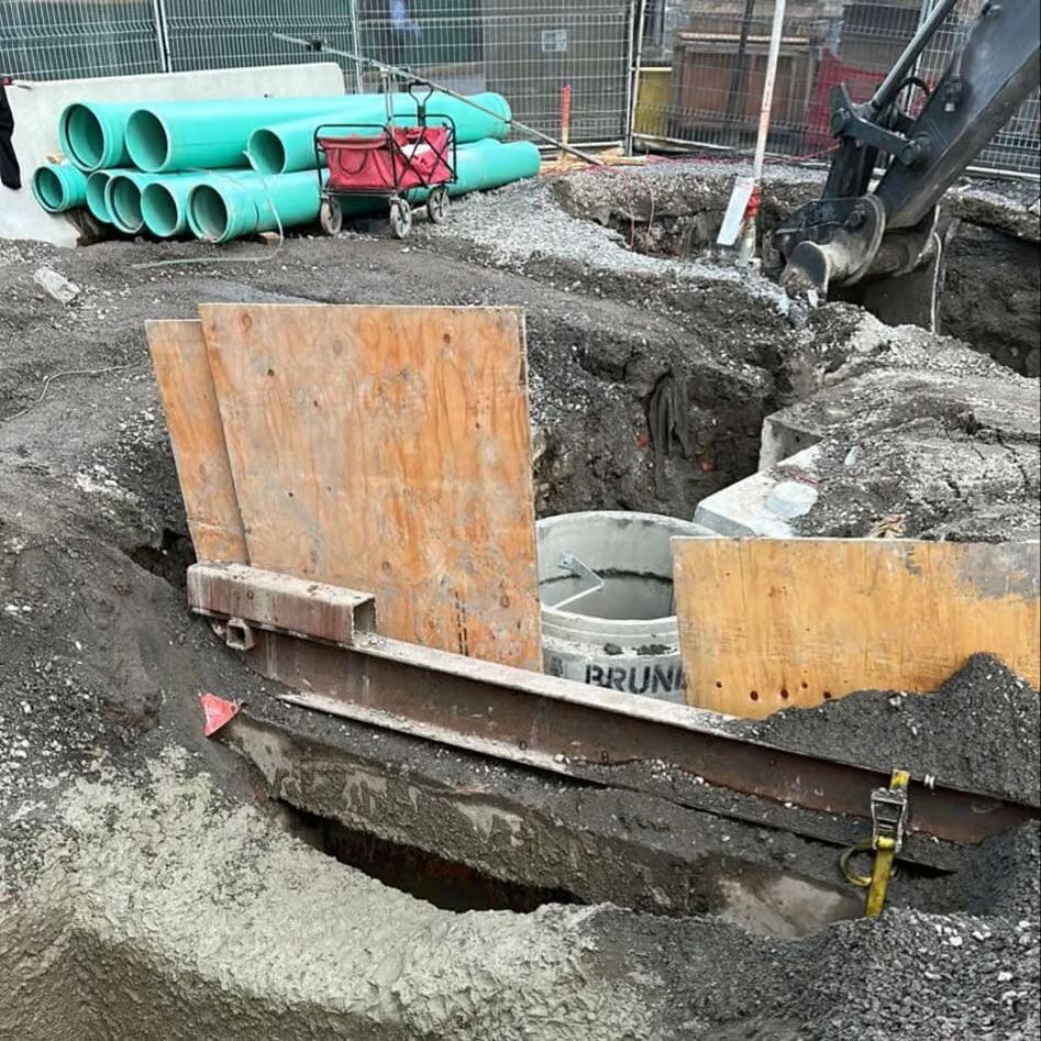 Chantier de construction avec tranchée et matériel d'excavation, planches de protection en bois et tuyaux.