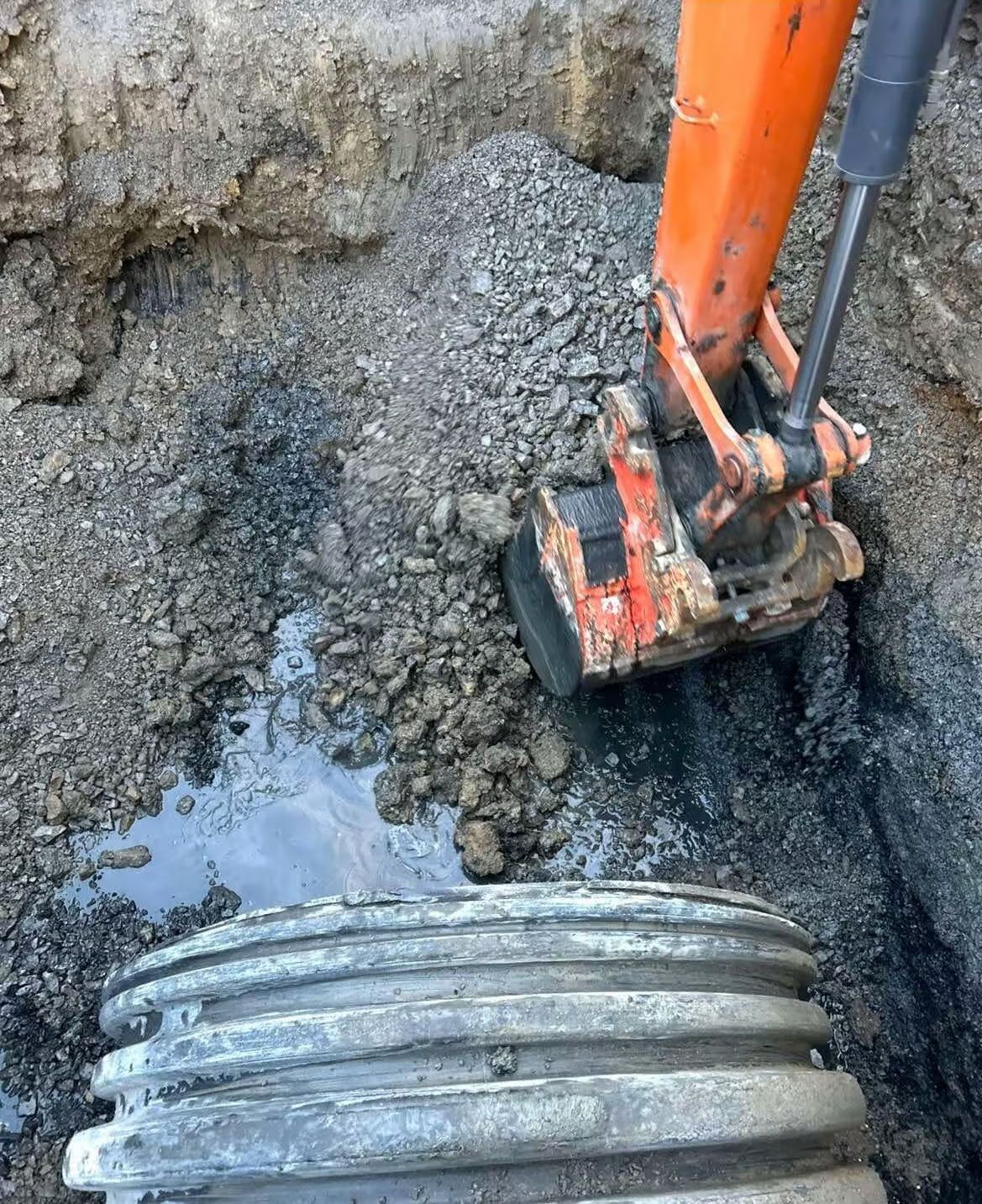 Une excavatrice travaille dans une tranchée à côté d'un tuyau ondulé. Le sol est sombre et humide.