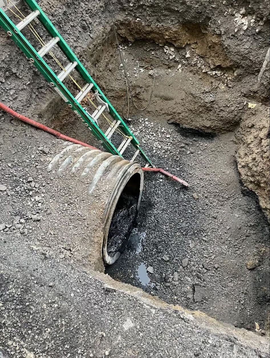 Un gros tuyau en béton dans une fosse d'excavation, avec une échelle verte et un tuyau rouge à proximité.