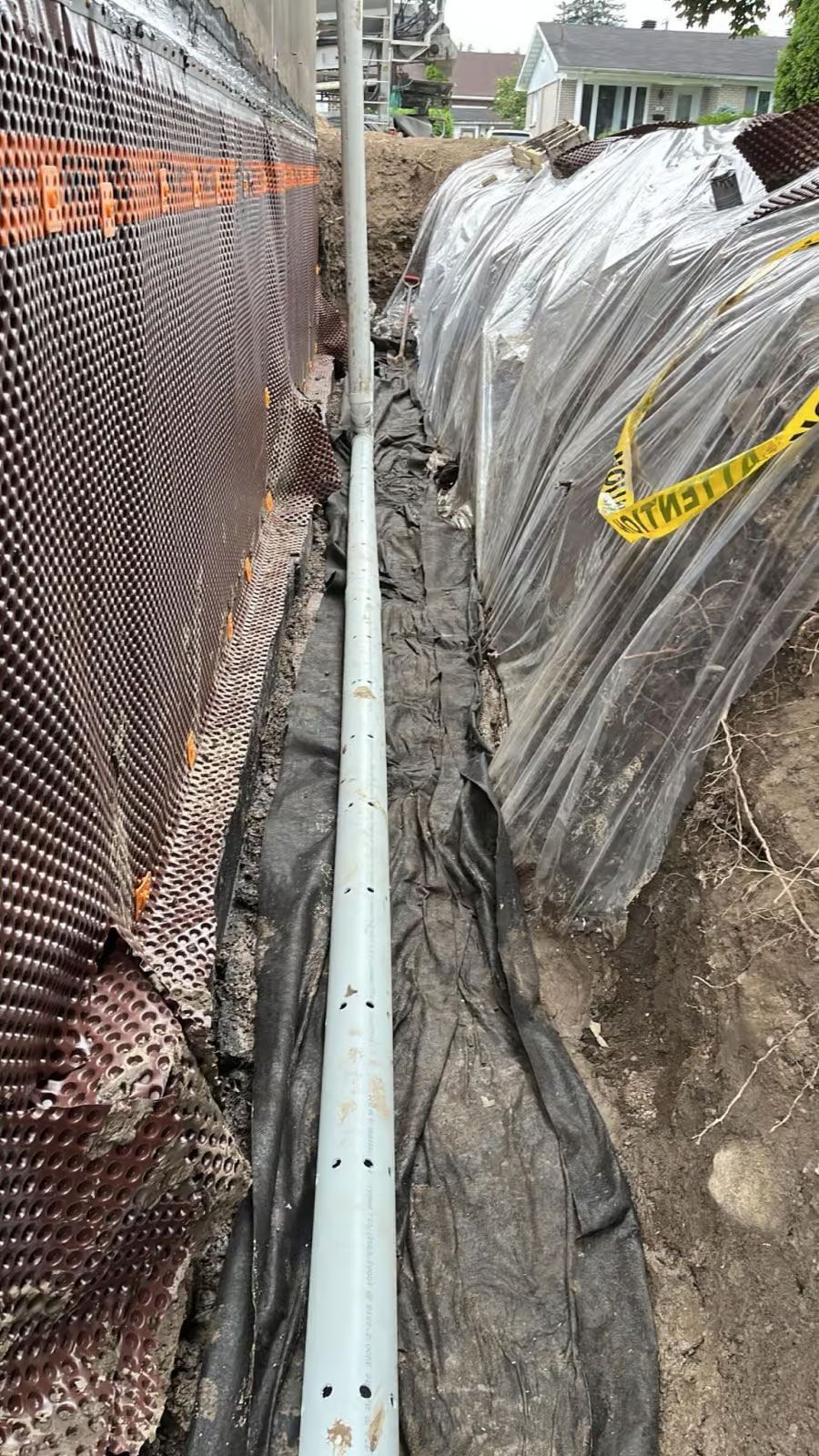 Tranchée avec tuyau perforé le long d'un mur de fondation, recouverte d'un tapis de drainage et enveloppée de plastique.