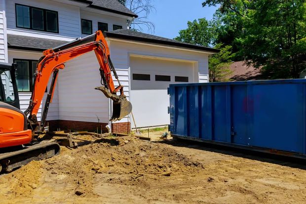 Une pelleteuse orange ramasse de la terre à côté d'une benne bleue, près d'un bâtiment blanc en construction.