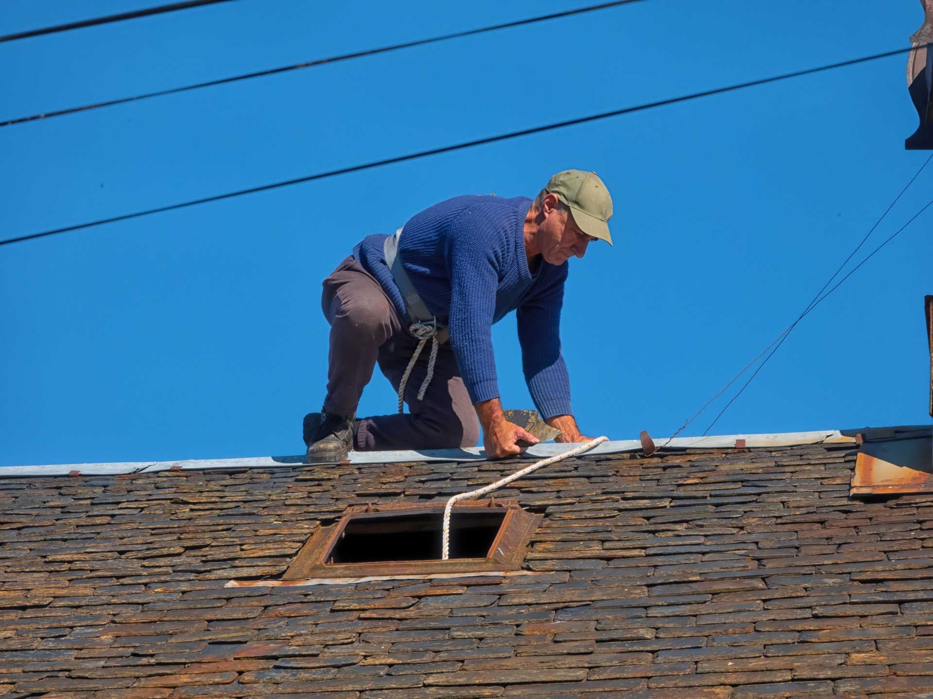 Worker repairing roof with safety harness, blue sky background, open roof hatch, power lines.