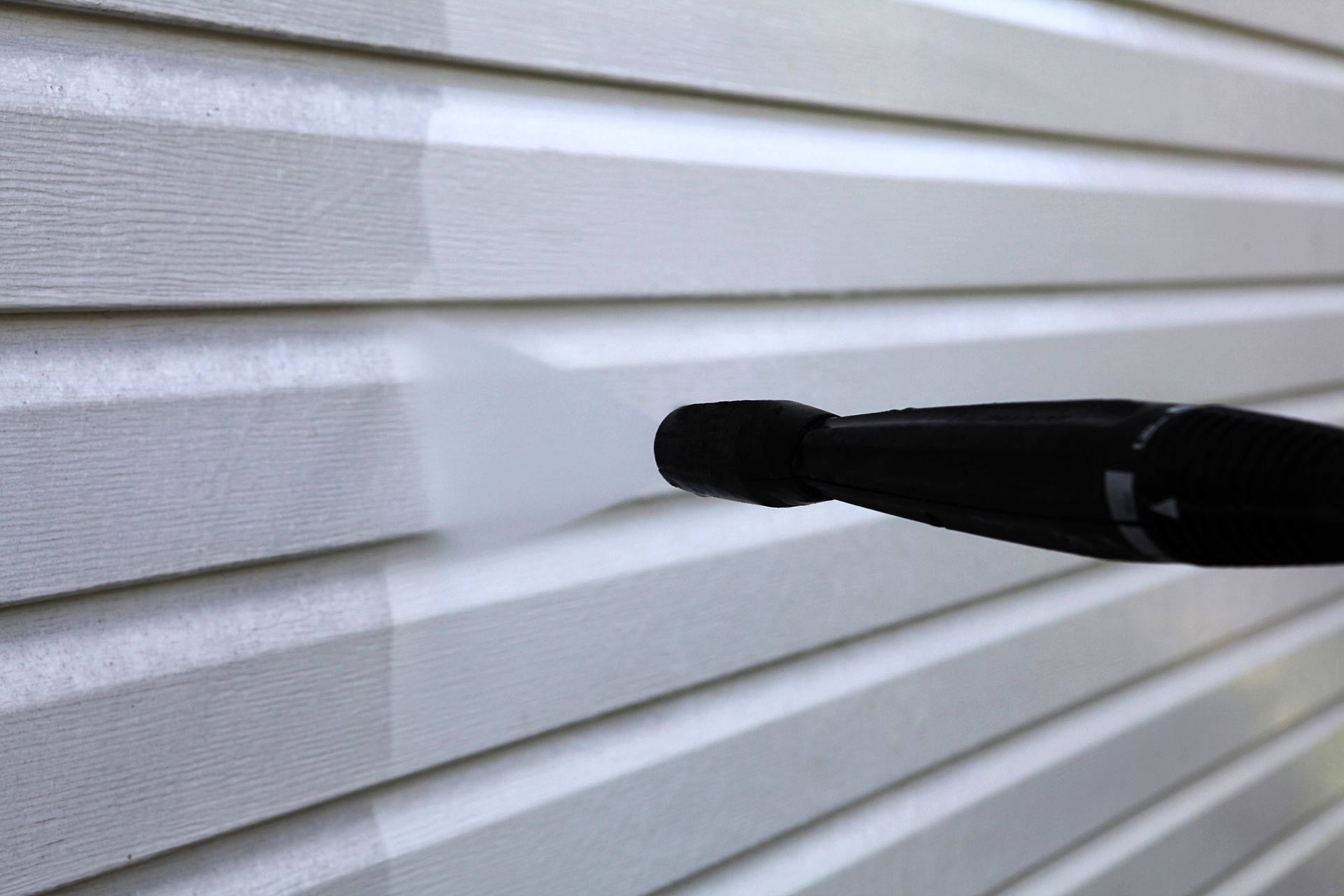 A person is cleaning a white siding with a high pressure washer.
