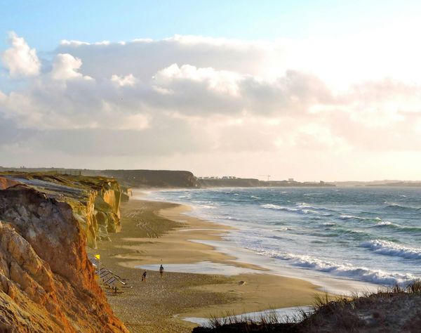 Acantilados rocosos junto al mar con vistas a una playa de arena y olas bajo un cielo nublado al atardecer.