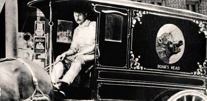 Man driving a Boar's Head wagon, with a boar's head emblem on the side. Black and white photo.