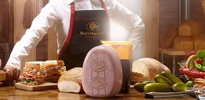 A deli worker stands behind a table of food. A large ham, sandwiches, bread, pickles, and peppers are displayed.
