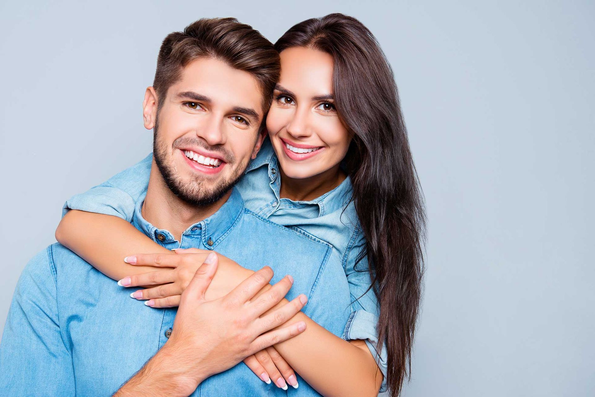 Lucent Orthodontics, San Antonio. A smiling couple in denim shirts posing against a light gray background, with the woman hugging the man from behind.