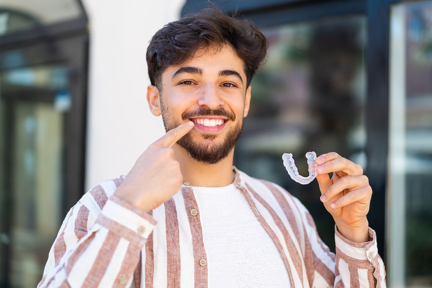 Smiling person wearing a striped shirt points to their teeth while holding a clear plastic dental aligner.