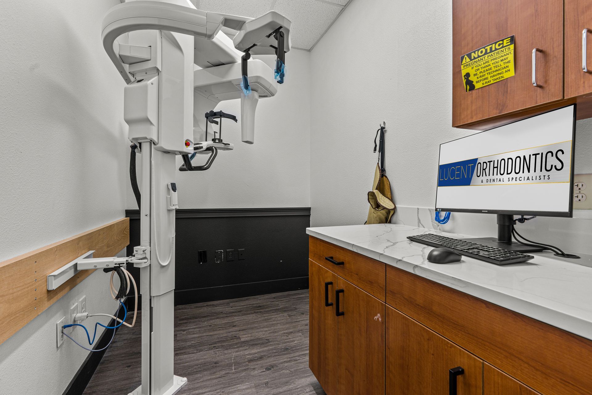 Lucent Orthodontics, San Antonio. A dental imaging room features a panoramic X-ray machine next to a computer station with a branded monitor on a wood desk.