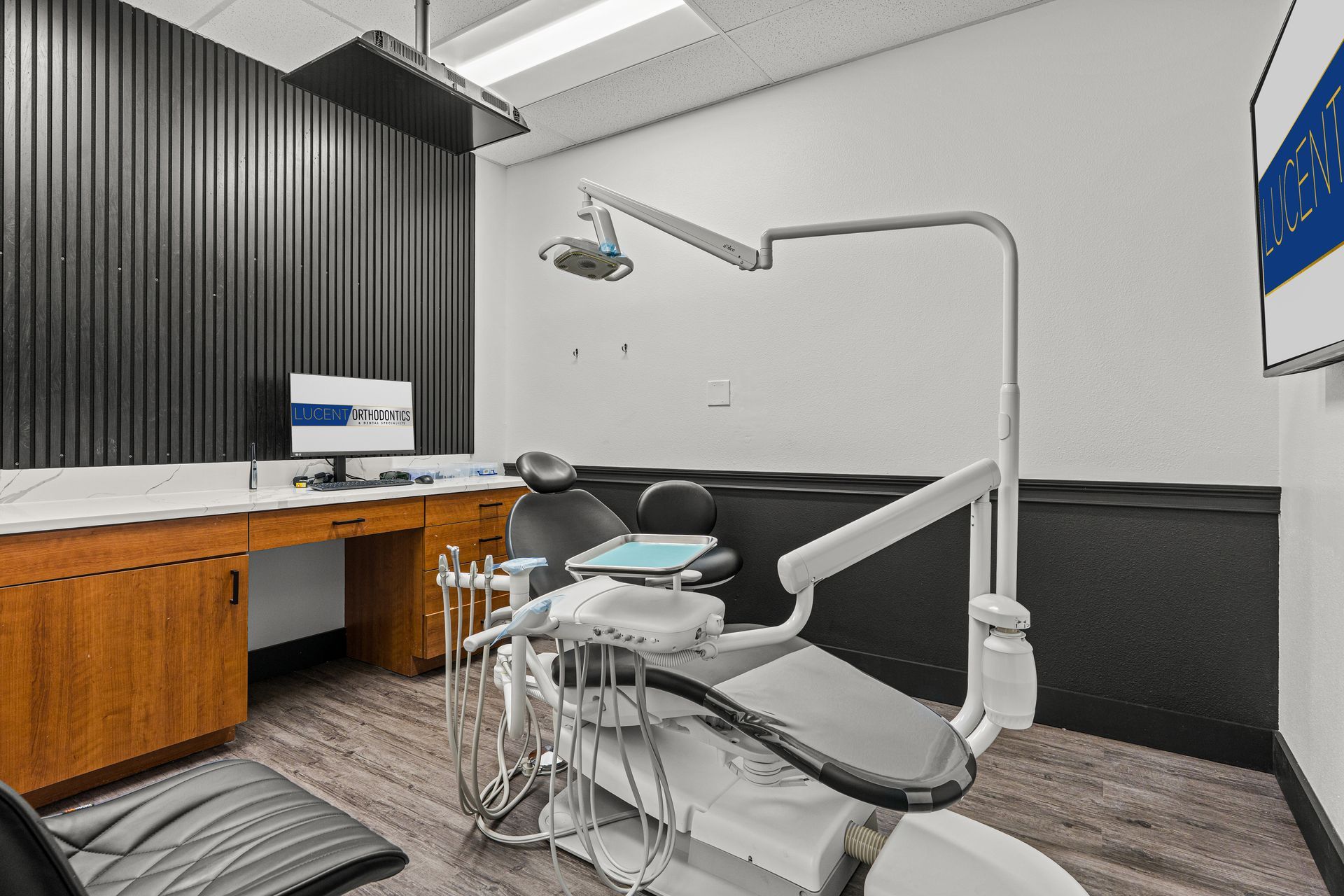 Lucent Orthodontics, San Antonio. A dental exam room with a reclined patient chair, dental equipment, and a wooden desk with a computer monitor.
