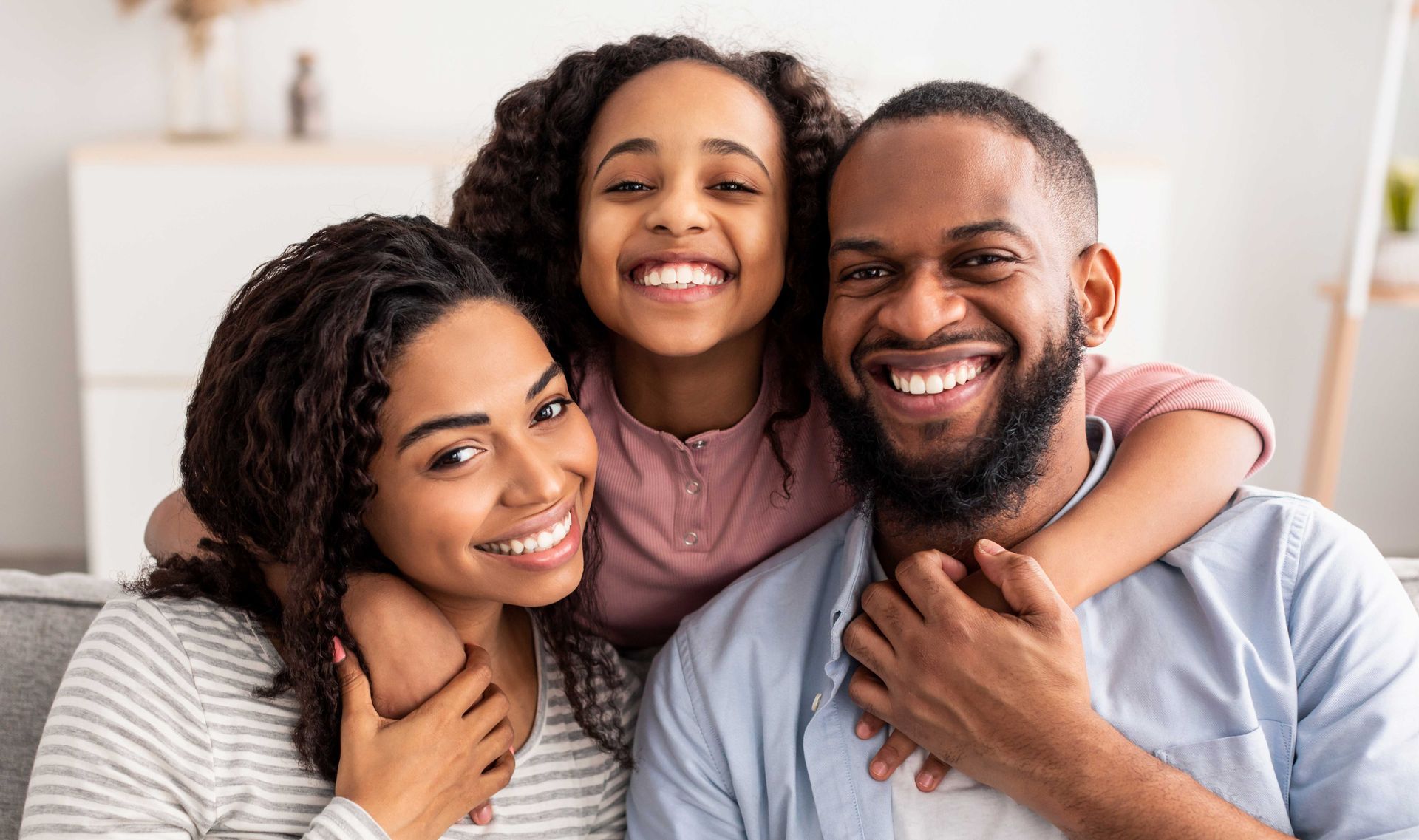 A smiling family of three huddled together on a couch, showing affection and warmth in a bright, indoor setting.