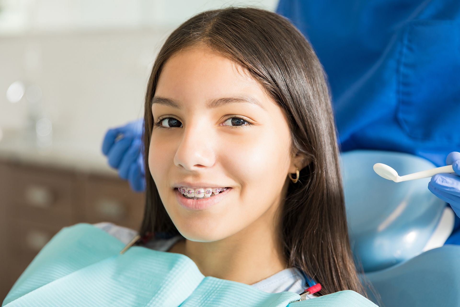 A person with braces sitting in a dental chair, facing a professional wearing blue medical scrubs and gloves.