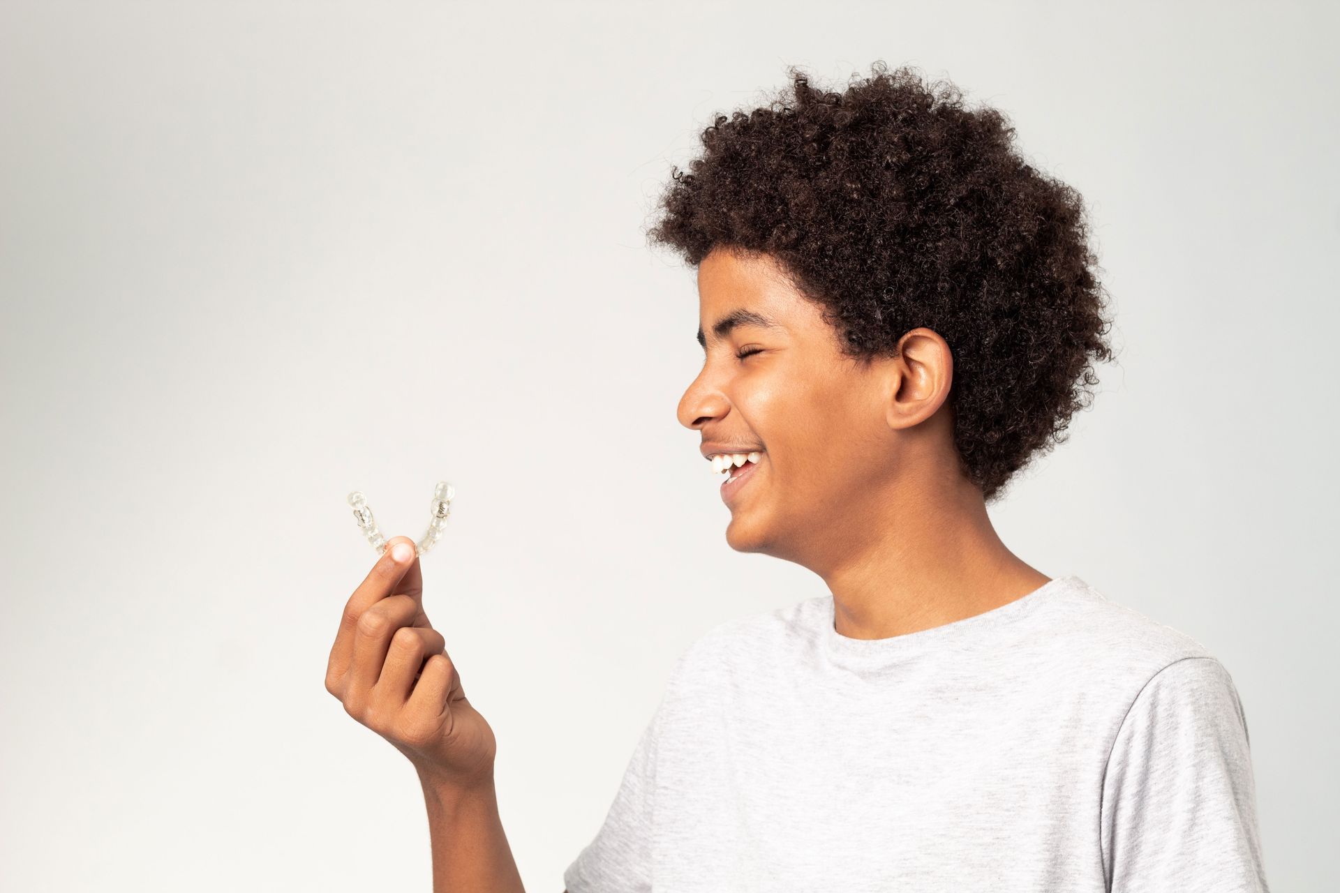 Lucent Orthodontics, San Antonio. A person in a light grey t-shirt smiles while holding a clear dental aligner in their hand against a white background.