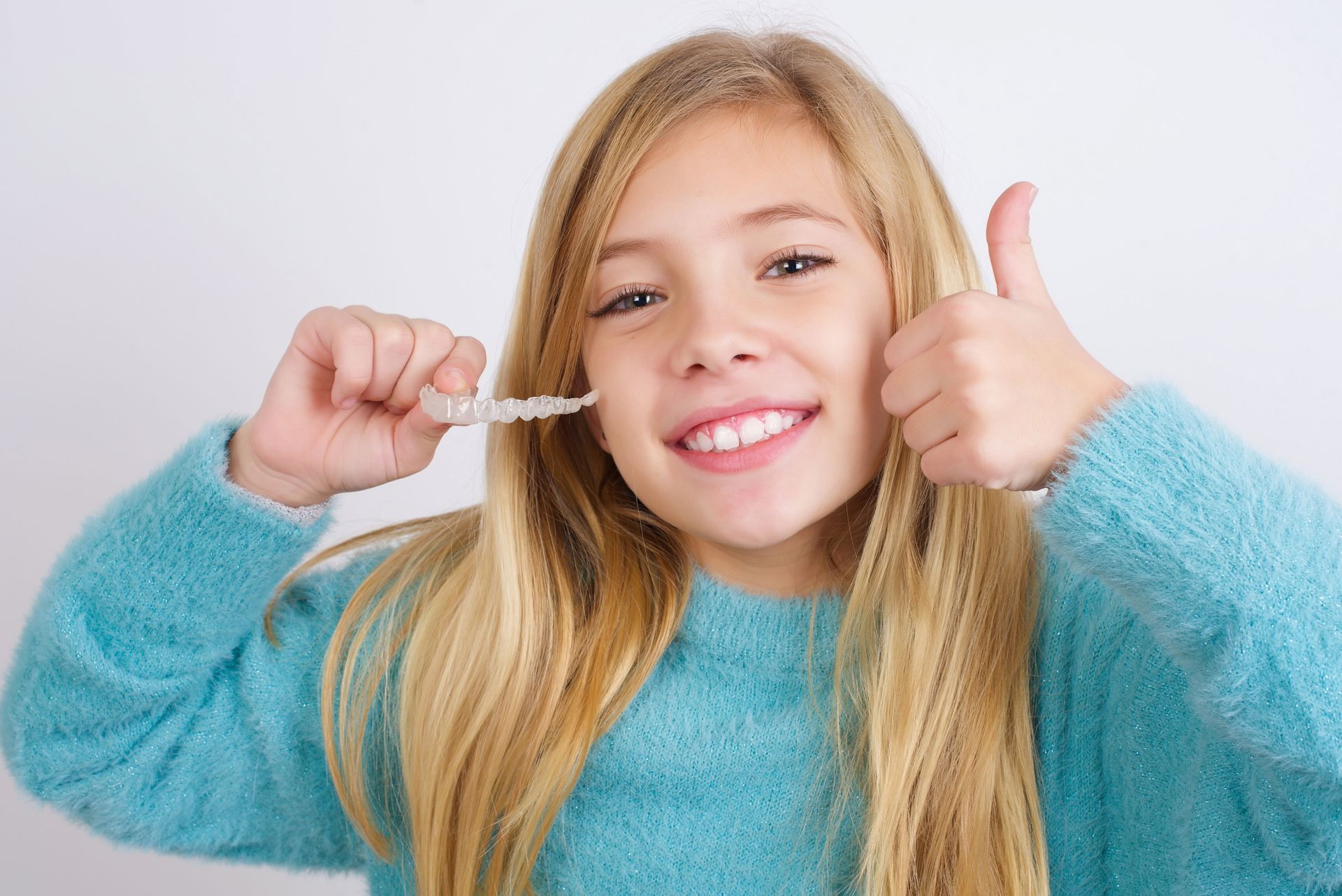 A person with blonde hair wearing a teal sweater smiles while holding a clear dental aligner and giving a thumbs up.