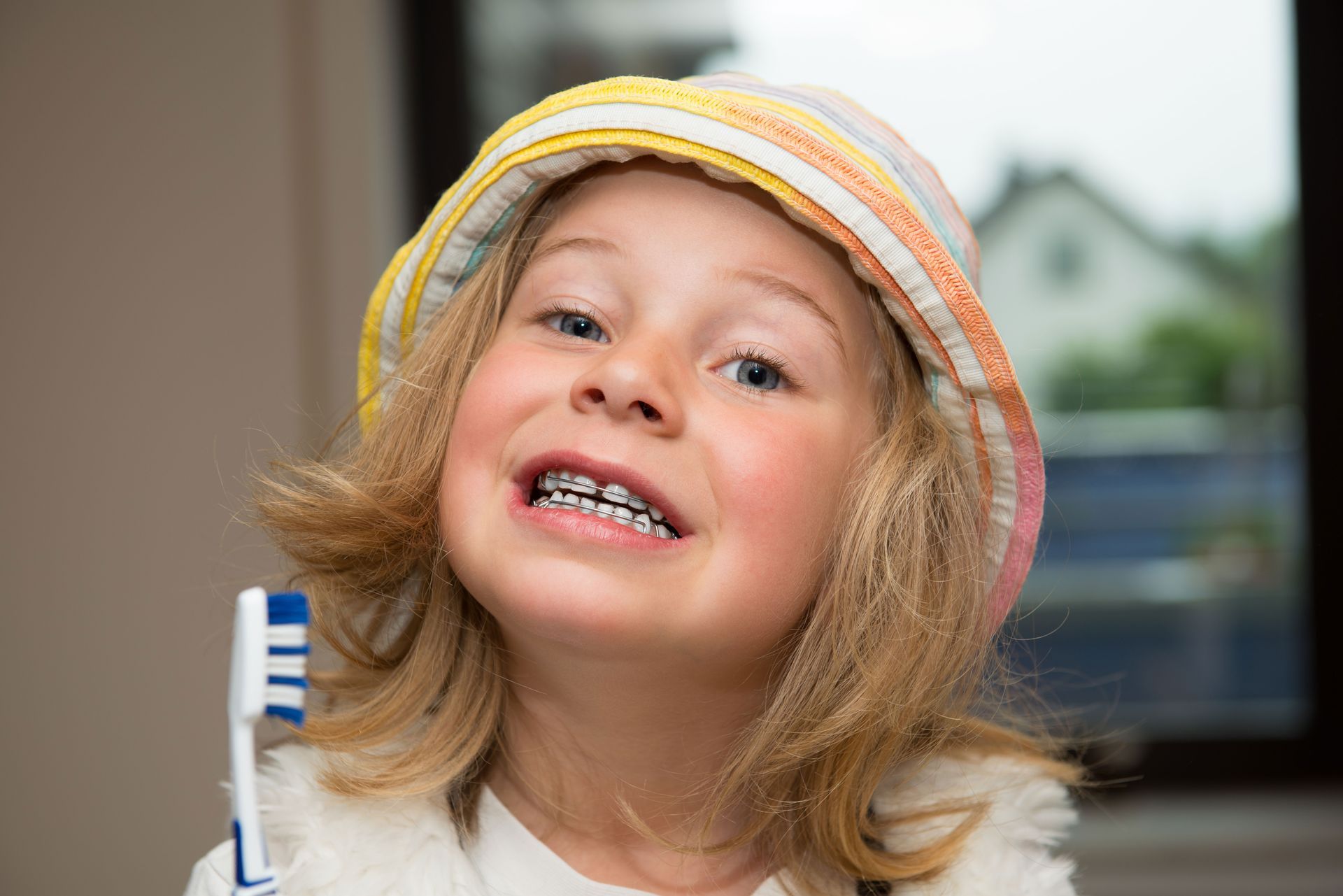 A person wearing a striped bucket hat and braces smiles while holding a toothbrush.