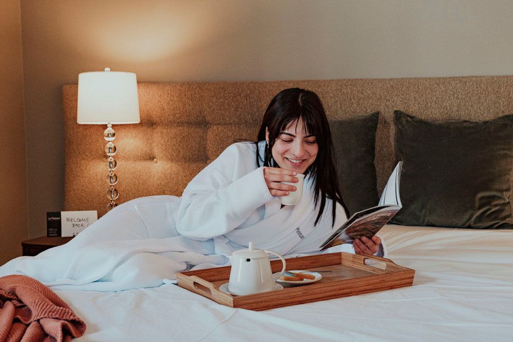 A woman is laying on a bed with a tray of food and drinks.