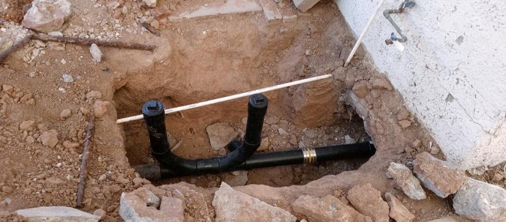 Black plumbing pipes with a brass fitting are in a dirt trench. A concrete structure is to the right.