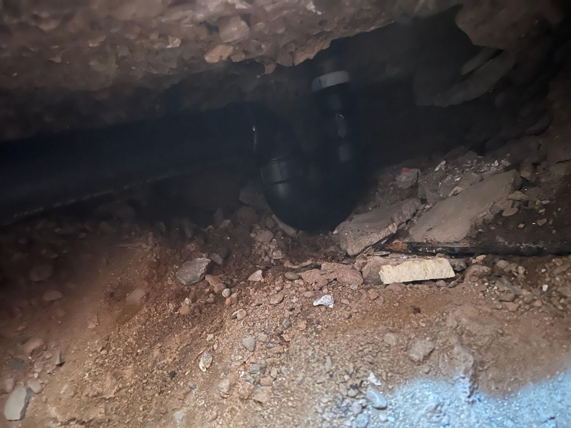 Looking into a hole in the ground, a black pipe with a curved connection is visible. Dirt and debris surround it.