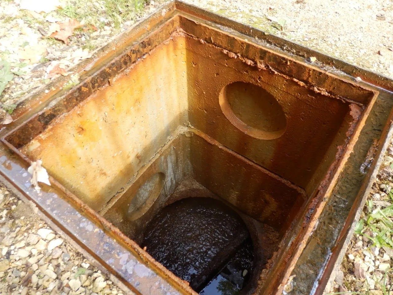 Open, rusty metal manhole with standing water and a dark, ball-shaped object inside.