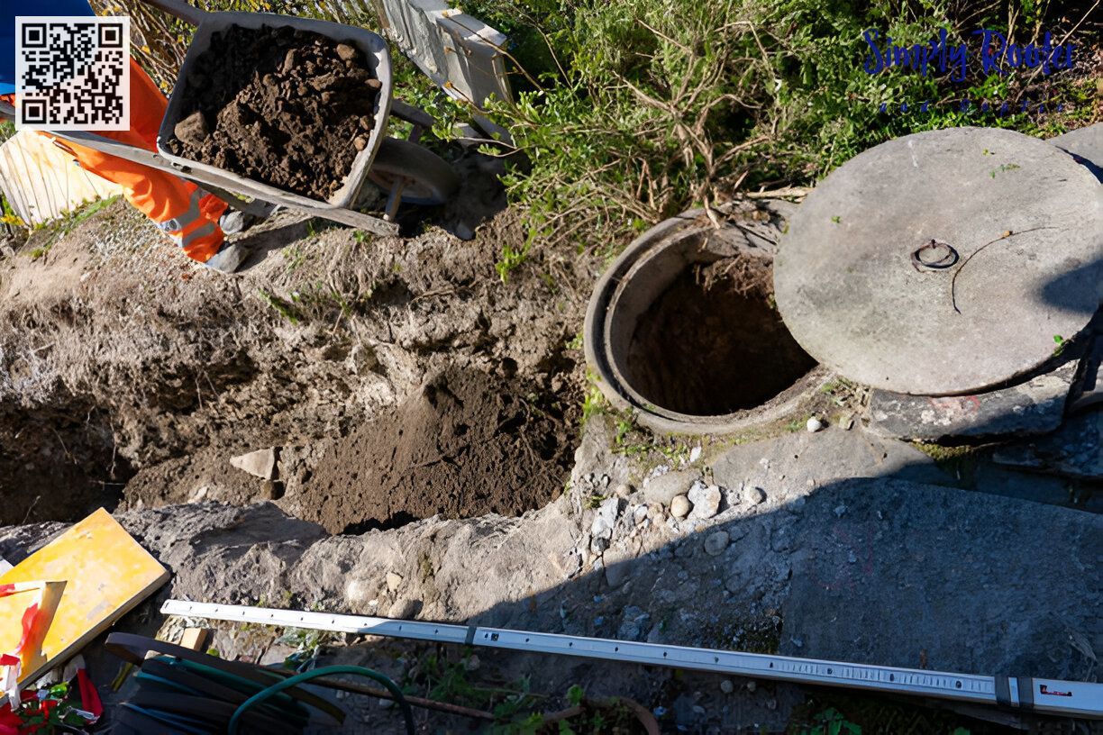 Open manhole with dirt surrounding it, tools, and a wheelbarrow filled with dirt.