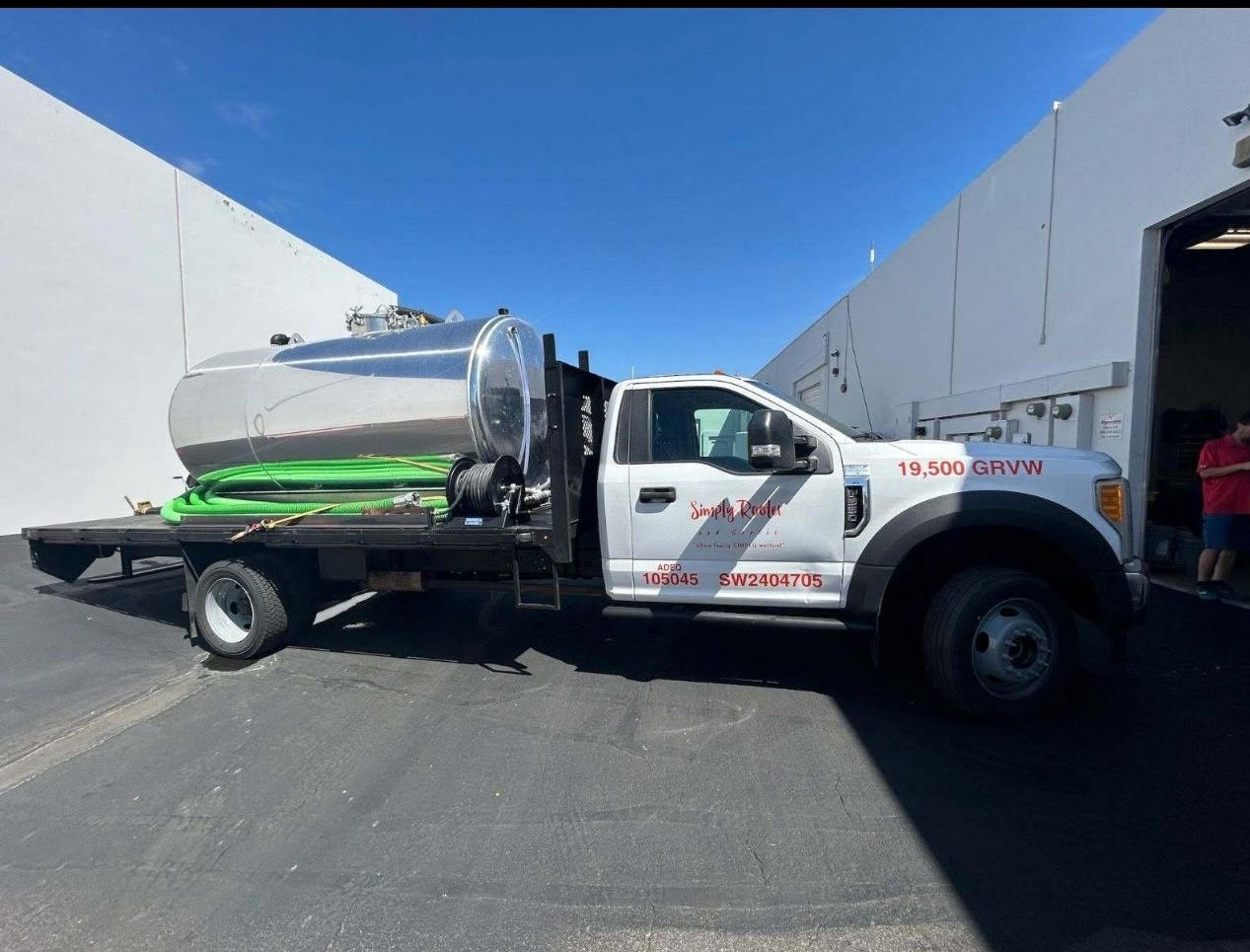 White truck with a silver tank on a flatbed, parked in front of a white building on a sunny day.
