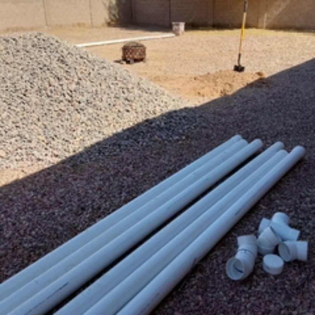 White PVC pipes and fittings on gravel, near a gravel pile and trench in an outdoor setting.