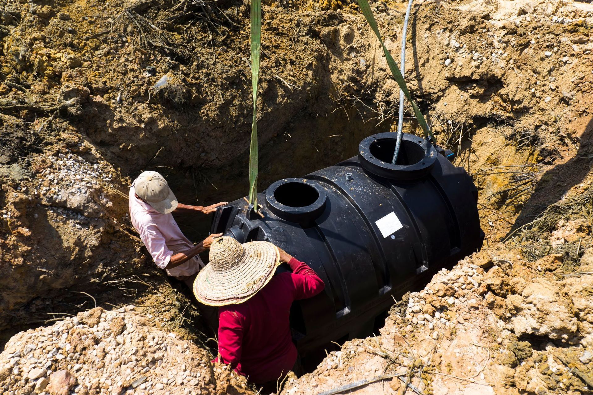 Two people lowering a black septic tank into a dug-out hole with ropes.