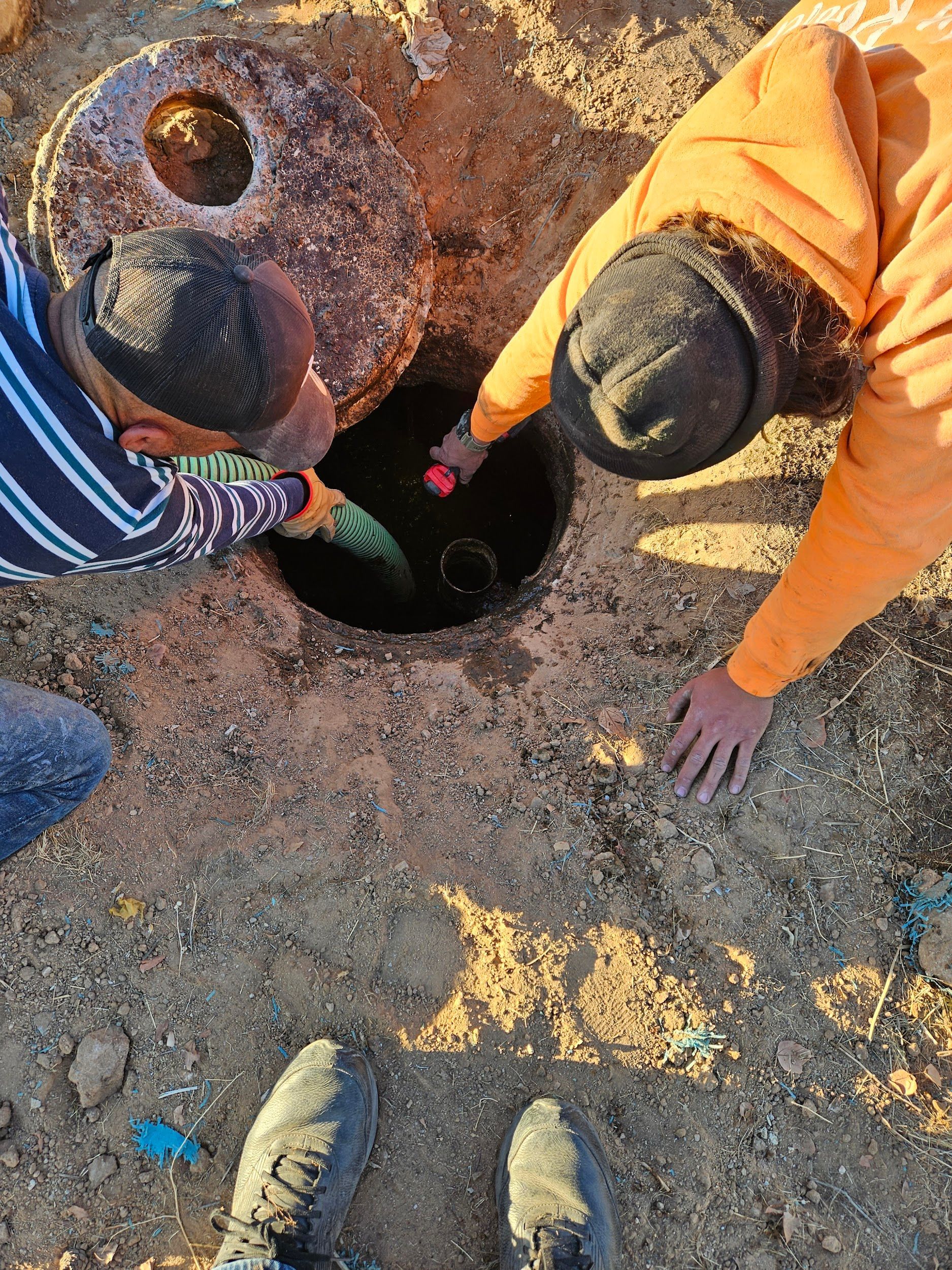 Two people inspecting a dark hole in the ground, a large metal cover nearby, on dirt.