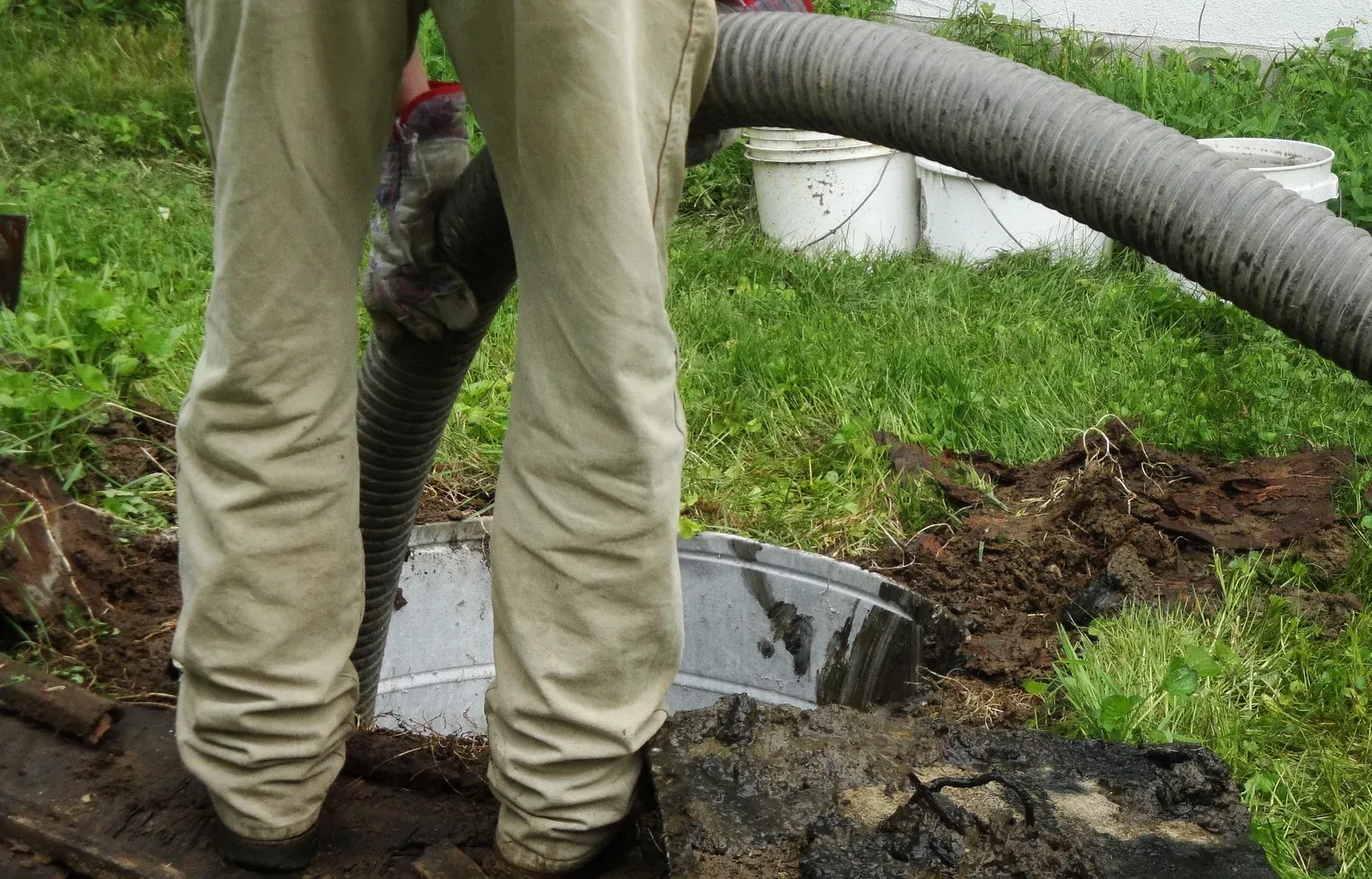 A person standing over an open septic tank, using a large suction hose to pump it out in a grassy outdoor area.