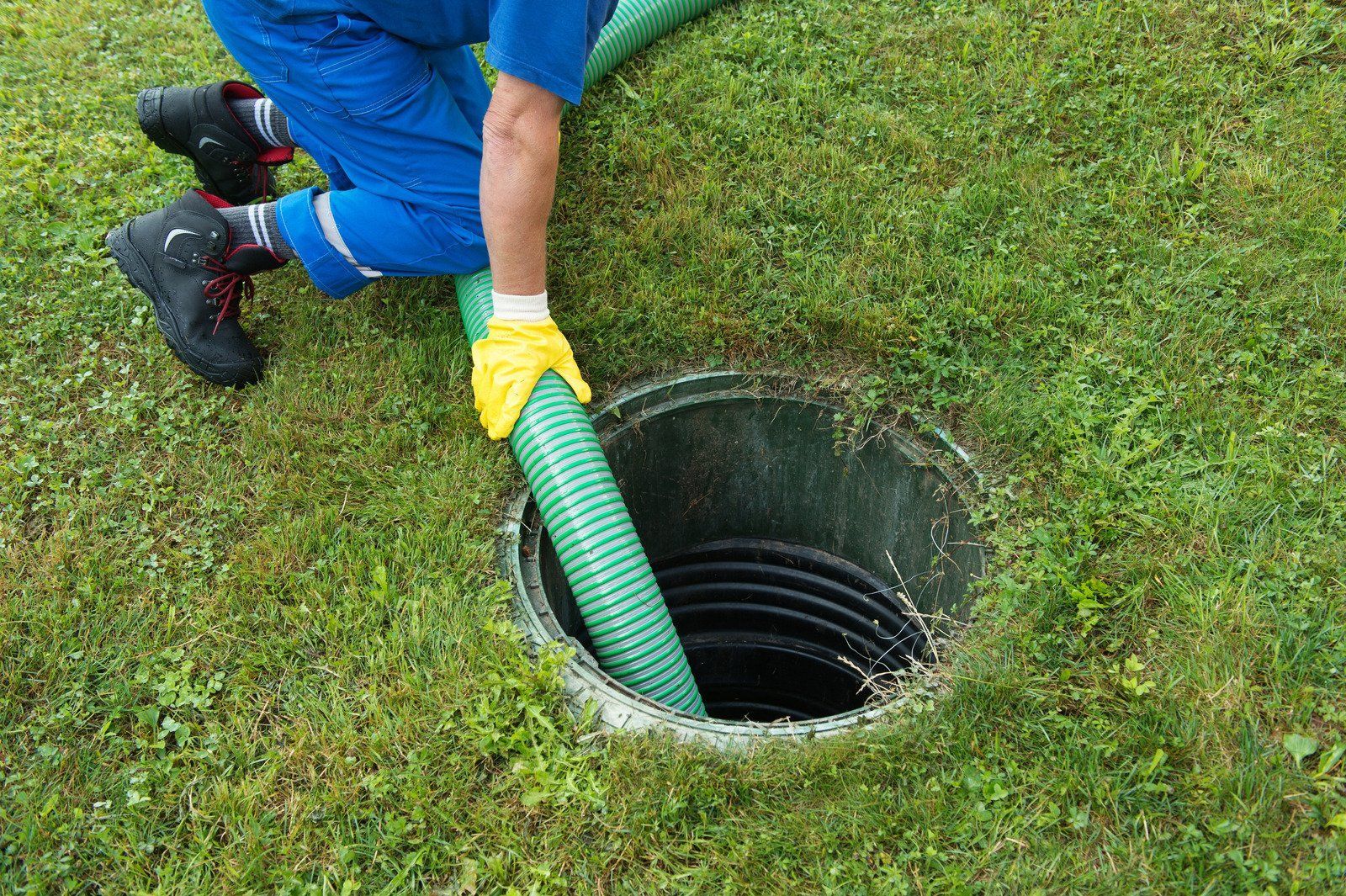 Worker in blue pants and yellow gloves cleaning a storm drain with a green hose on wet grass