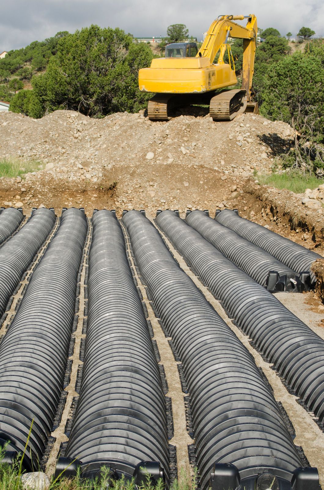 Yellow excavator on a dirt pile above a trench containing multiple black septic chambers.