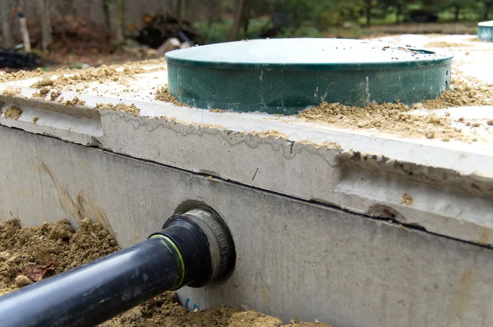 Concrete septic tank with green lid and black pipe connection, partially buried in soil.