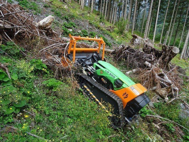 Green and orange tracked forestry robot on a steep, grassy hillside clearing debris near trees.