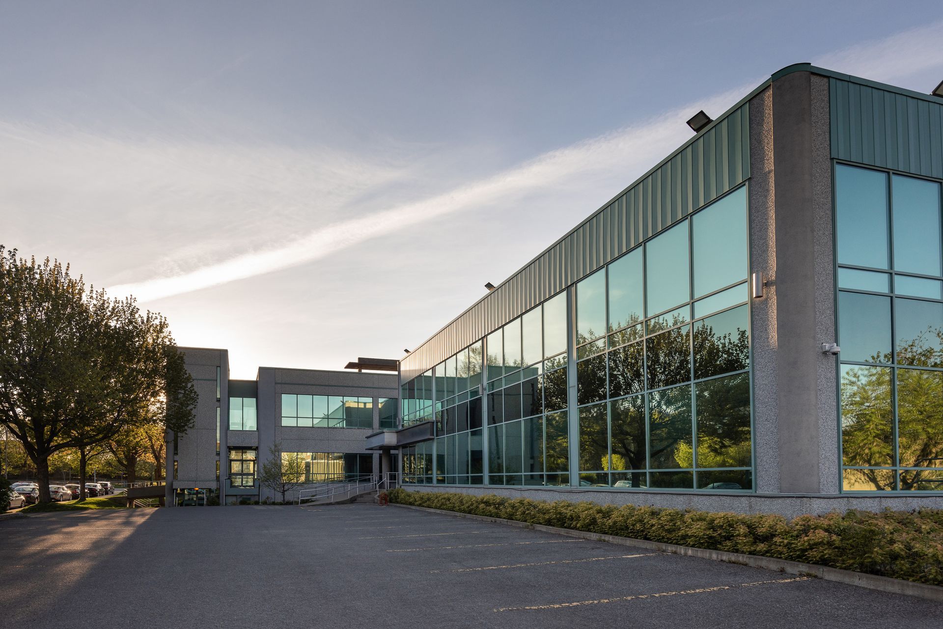 Modern office building with large glass windows reflecting the sky, set in a parking lot.