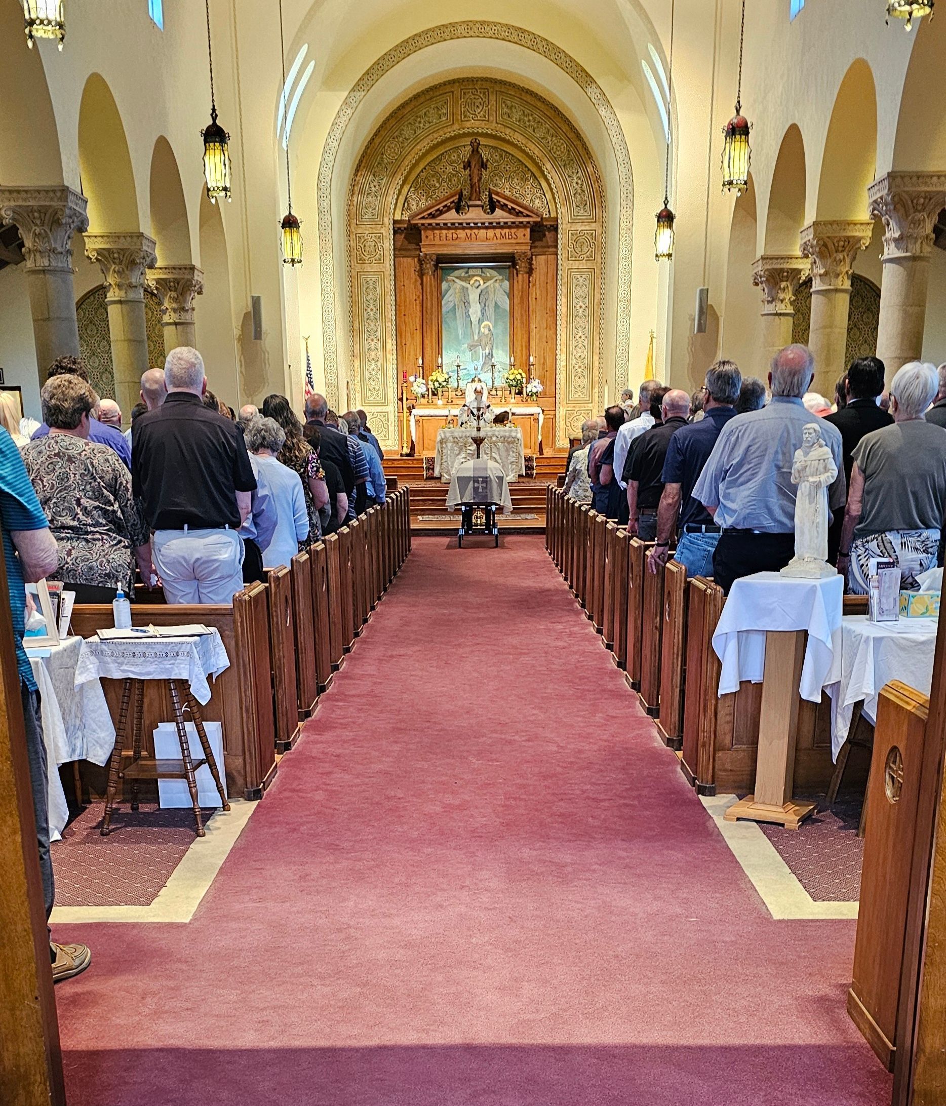 Interior of a church during a service, people seated in pews, facing altar. A priest stands at the altar.