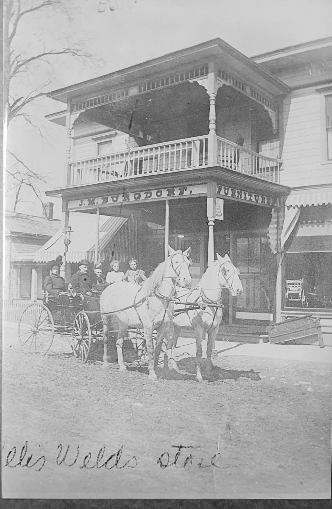Horse-drawn carriage in front of a two-story building with a decorated porch and balcony. People are in the carriage.
