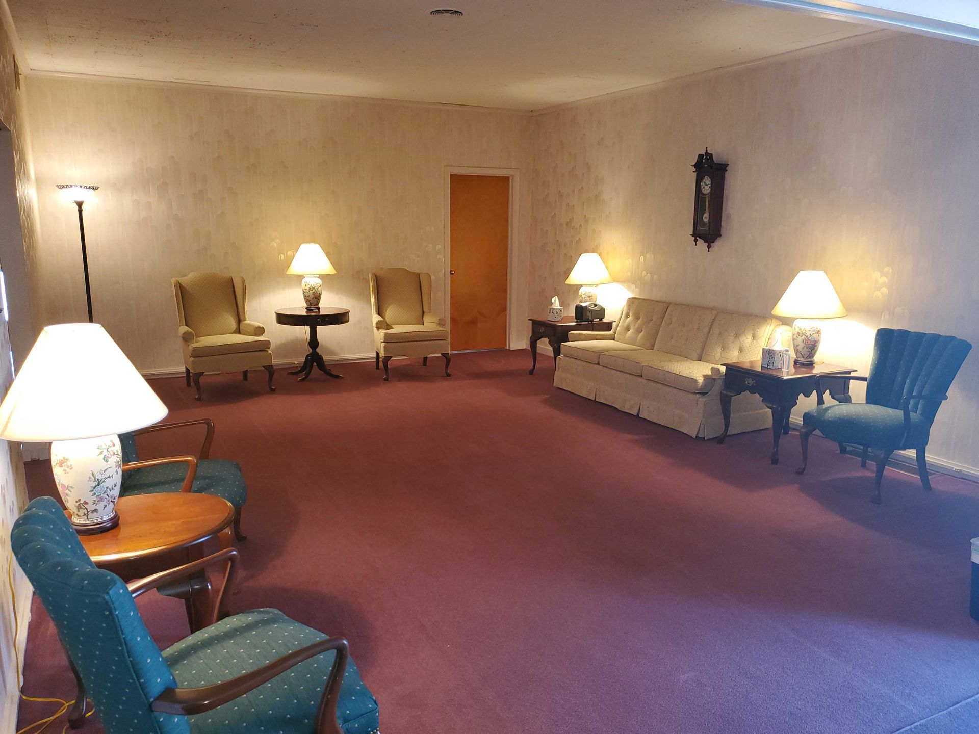 A room with pink carpet, beige walls, and various upholstered chairs, lamps, and a sofa.