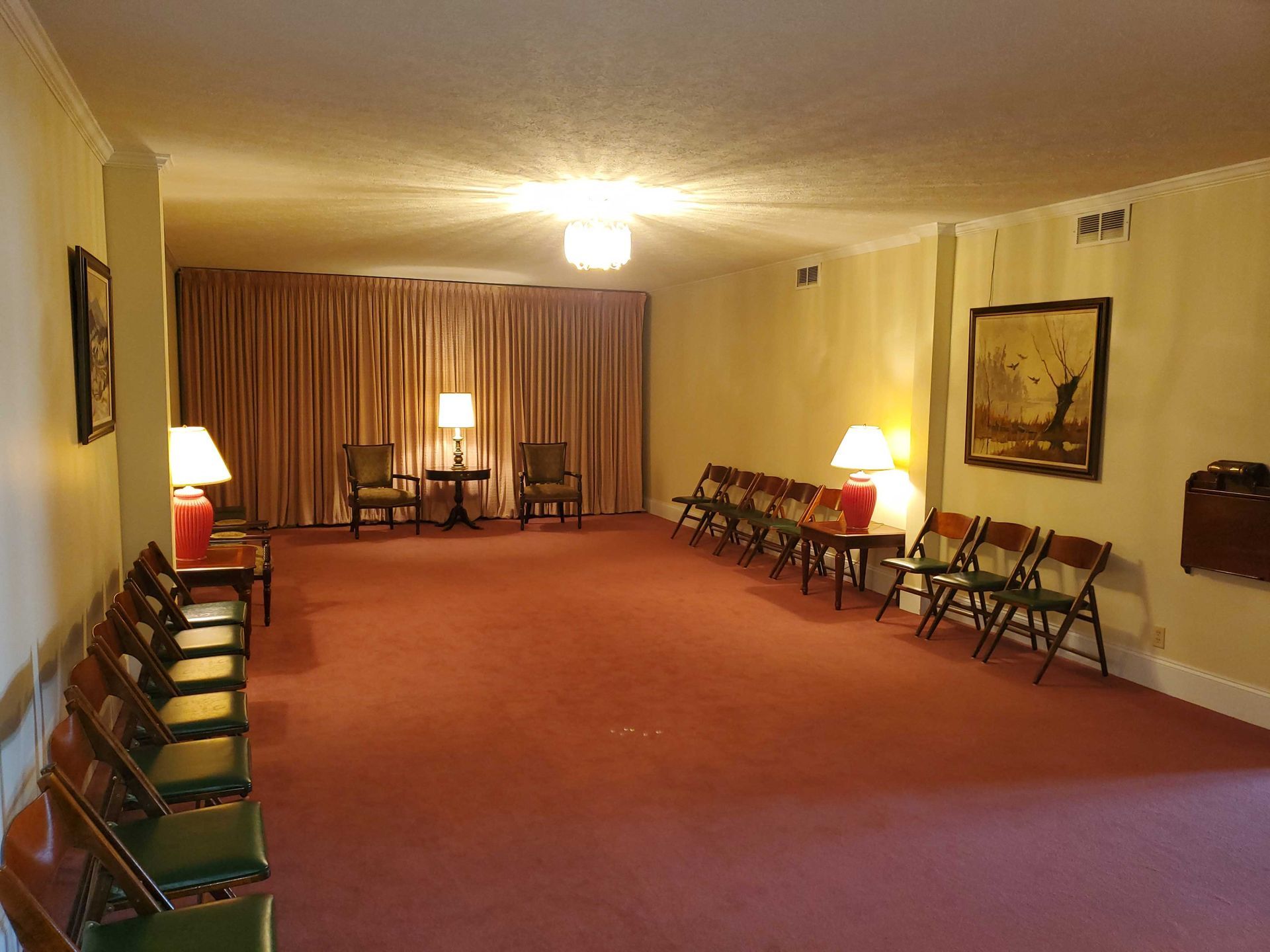 Interior of a dimly lit funeral home parlor with chairs, lamps, red carpet, and a large curtained area.