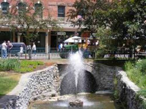 Fountain in a stone-lined waterway, downtown setting. People walking, buildings in the background.