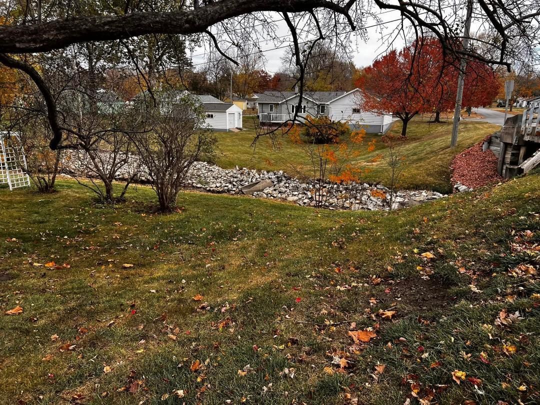Grassy landscape with a creek bed and fall foliage, including red and orange trees, on an overcast day.